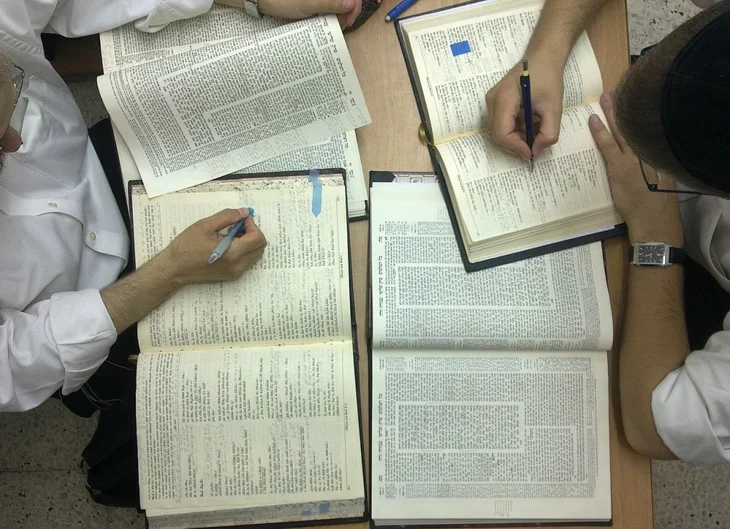 Four open Jewish books on a table with men writing notes